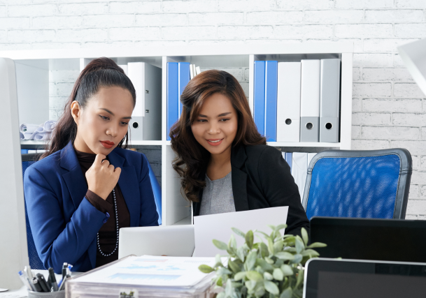 two-asian-female-colleagues-sitting-together-office-looking-document 1